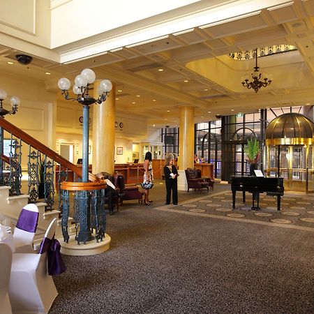 Stamford Grand Adelaide Hotel Glenelg Interior photo The photo shows a luxurious interior of a hotel or a restaurant lobby. There is an elegant staircase with ornate railings leading to an upper level. The space features high ceilings with decorative chandeliers and ample natural light coming through l