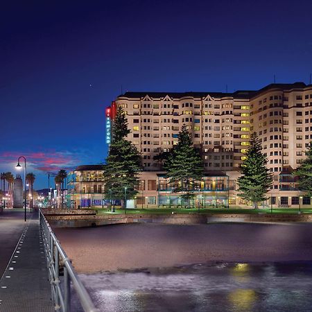Stamford Grand Adelaide Hotel Glenelg Exterior photo The photo shows a waterfront scene at dusk, featuring a large building lit up against a darkening sky. The architecture appears modern and prominent, with several windows illuminated. In the foreground, there is a walkway or pier with fencing along t