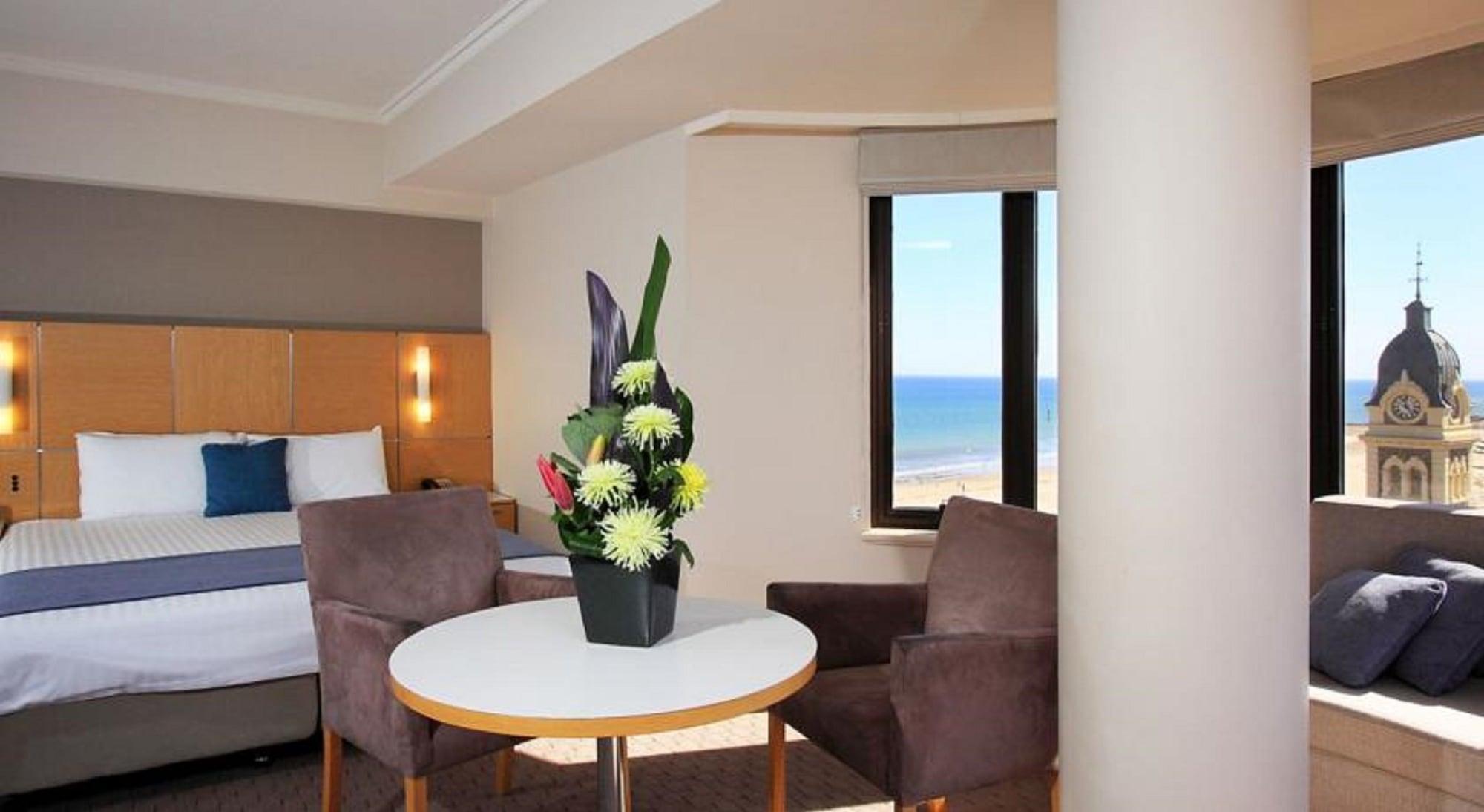 Stamford Grand Adelaide Hotel Glenelg Exterior photo The photo shows a cozy hotel room with a comfortable seating arrangement. There are two brown chairs positioned around a round table, which features a decorative flower arrangement in a black vase. The background includes a window that offers a view 