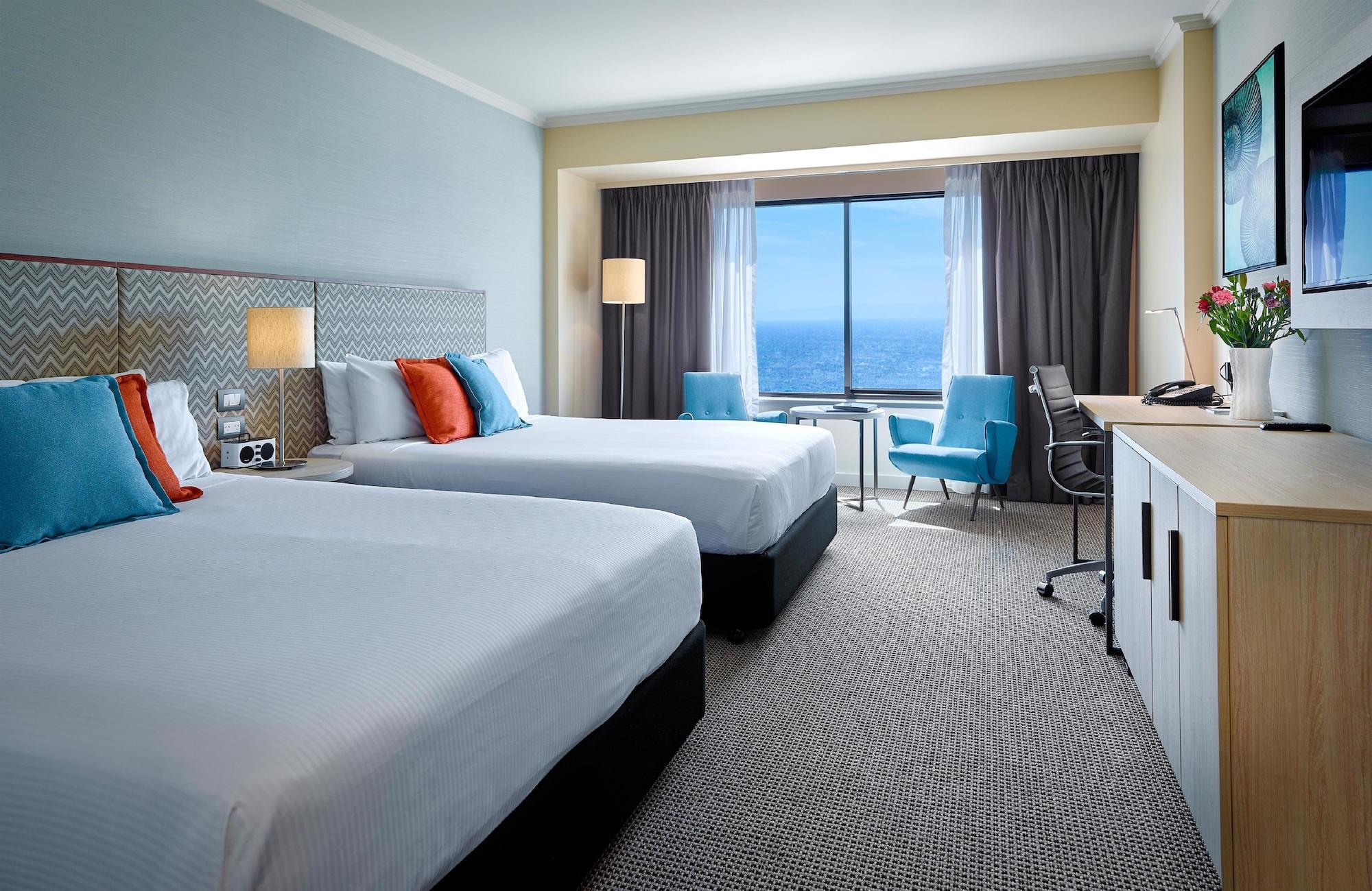 Stamford Grand Adelaide Hotel Glenelg Exterior photo The photo shows a well-decorated hotel room featuring two queen-sized beds with white linens and colorful accent pillows. There is a large window with curtains that offers a view of the ocean. A small seating area with a blue chair and a matching des