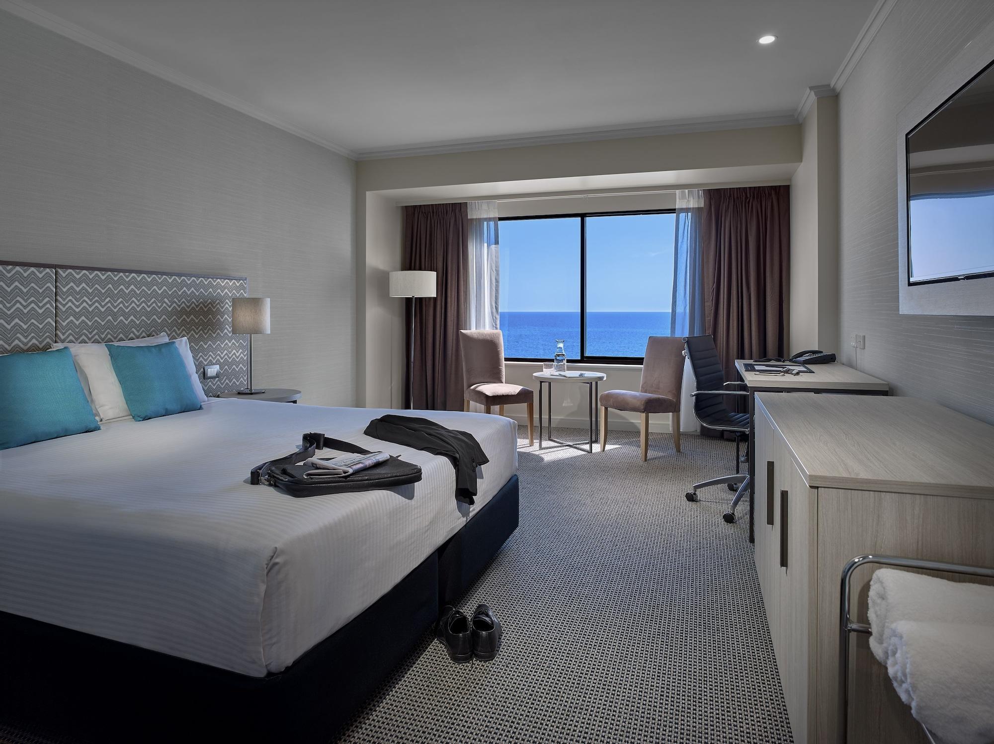 Stamford Grand Adelaide Hotel Glenelg Exterior photo The photo shows a modern hotel room featuring a queen-sized bed with white linens and two aqua blue decorative pillows. There are bedside lamps and a large window with a view of the ocean. The room also has a small seating area with two chairs and a 