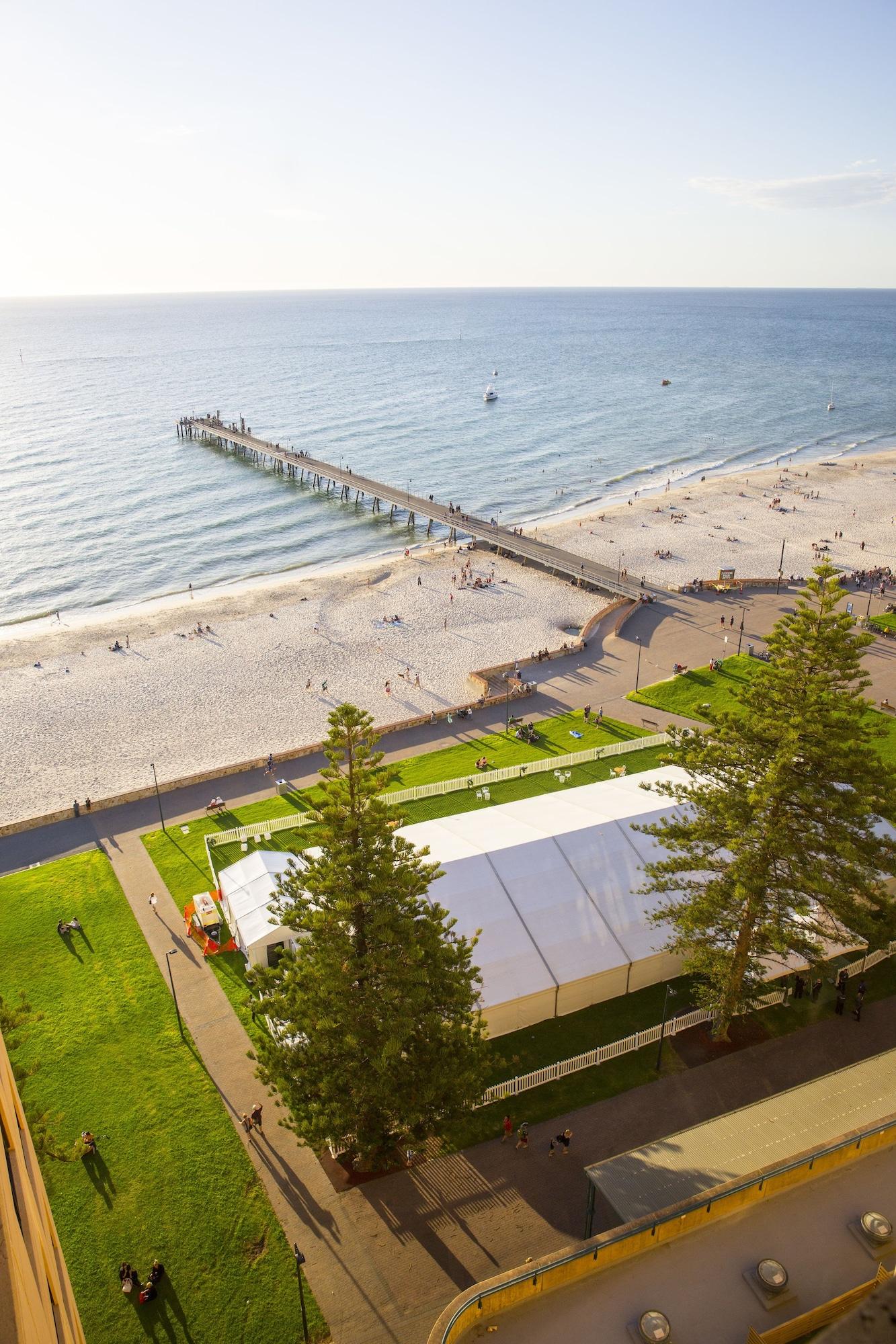 Stamford Grand Adelaide Hotel Glenelg Exterior photo The photo shows a scenic view of a beach with a long pier extending into the water. The beach is sandy, and there are people enjoying the sun and walking along the shore. In the foreground, there is a large white tent set up on a grassy area, likely 