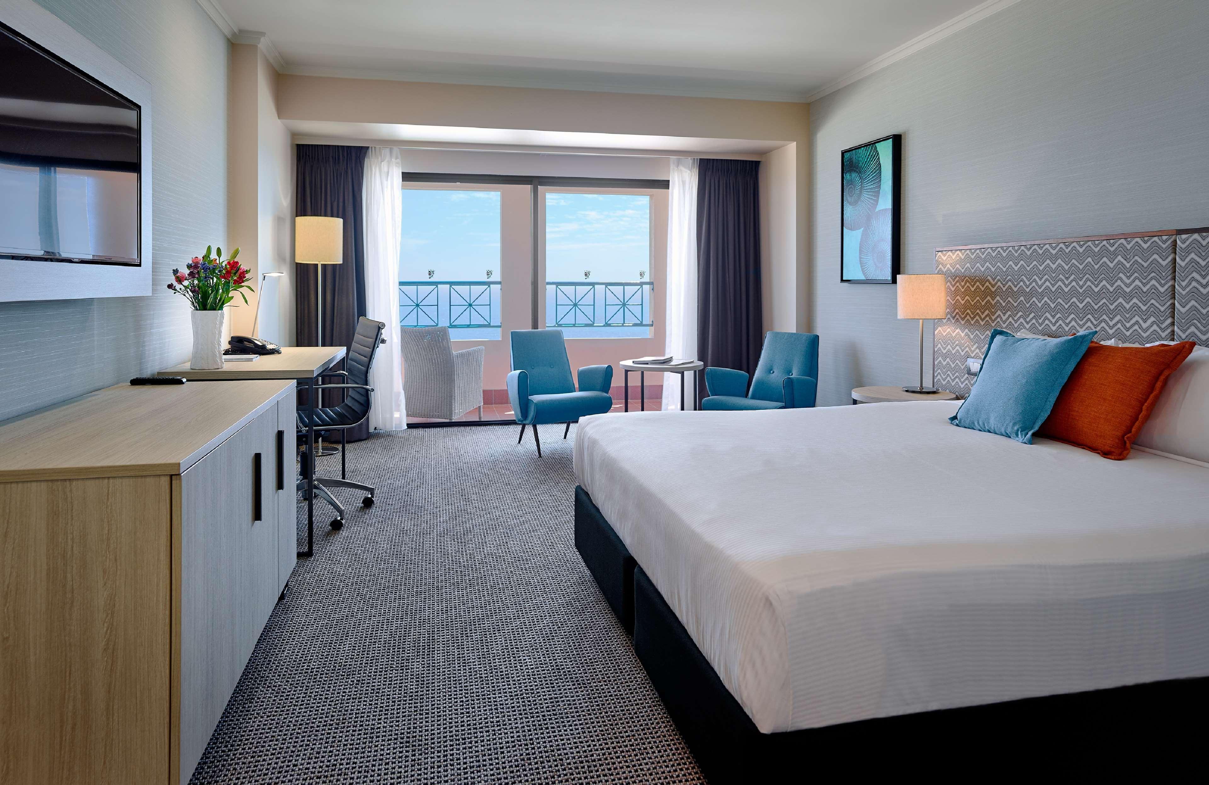 Stamford Grand Adelaide Hotel Glenelg Exterior photo The photo depicts a modern hotel room. The room features a large bed with a white comforter, and there are decorative pillows in orange and blue. There is a flat-screen TV mounted on the wall, and a desk with an office chair is positioned near the wi