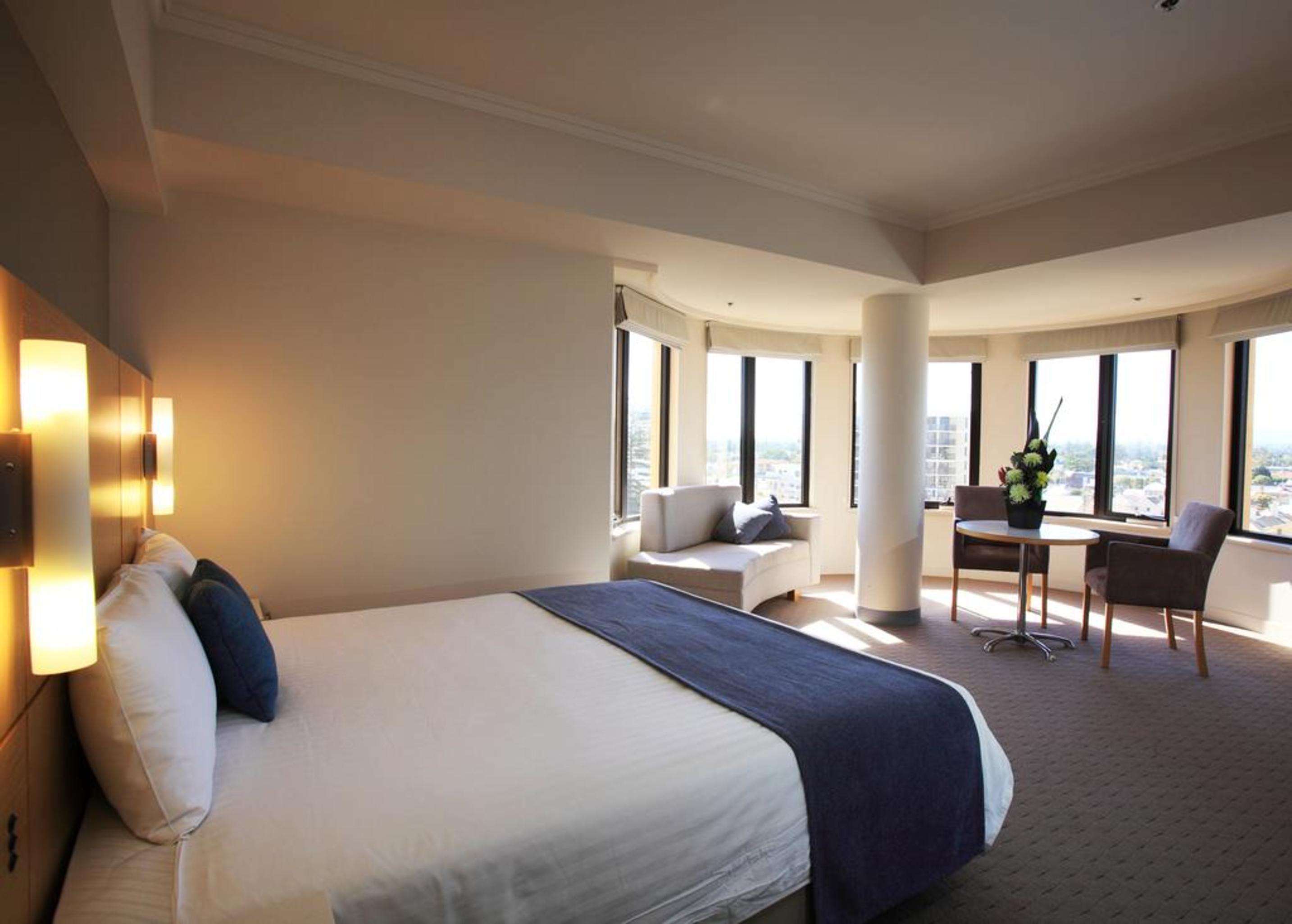 Stamford Grand Adelaide Hotel Glenelg Exterior photo The photo shows a hotel room with a modern and spacious design. There is a large bed with white linens and a dark blue throw blanket. The room features multiple windows that allow plenty of natural light, and there is a small seating area near the wi