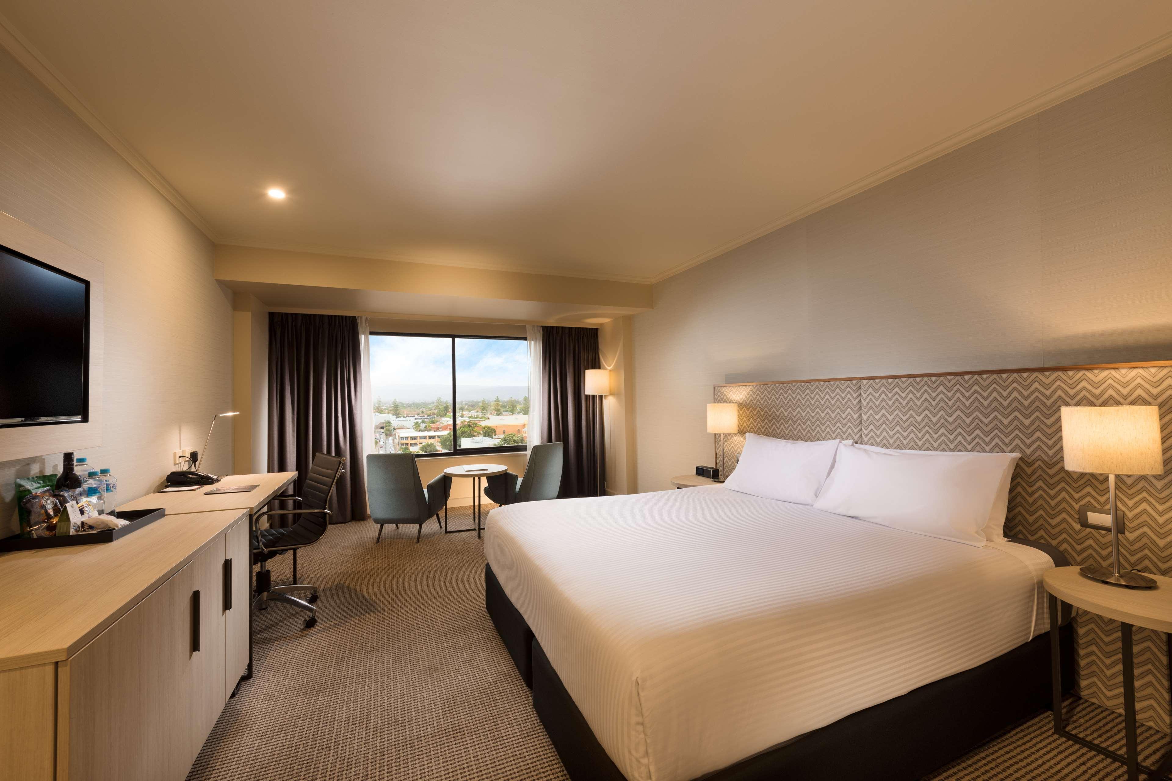 Stamford Grand Adelaide Hotel Glenelg Exterior photo The photo shows a modern hotel room. It features a large bed with white linens, a desk with a chair, and a flat-screen TV mounted on the wall. There are two armchairs and a small table near a large window that lets in natural light, providing a view 