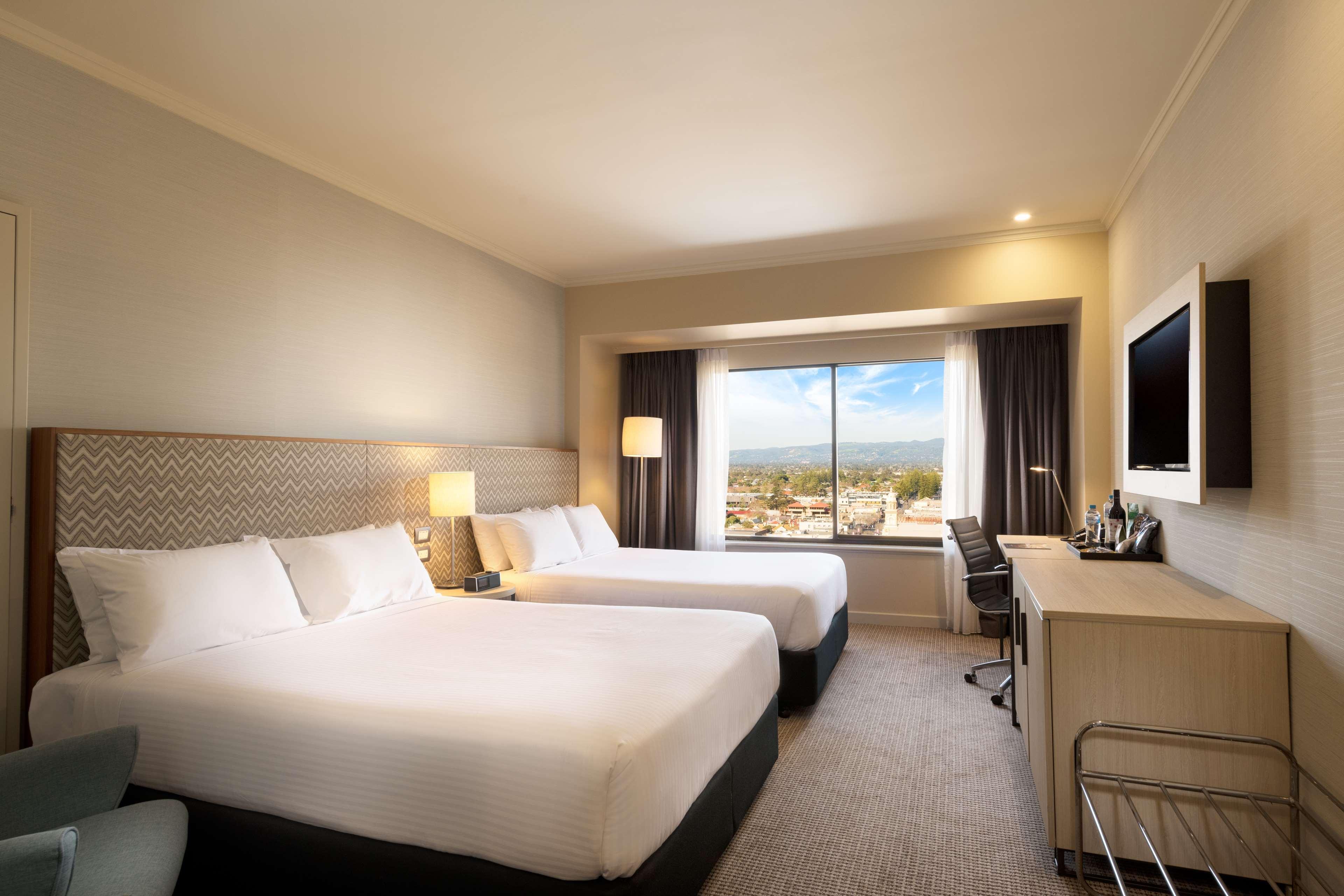 Stamford Grand Adelaide Hotel Glenelg Exterior photo The photo shows a modern hotel room featuring two beds with white linens. There are bedside tables with lamps next to each bed. A large window provides natural light and offers a view of the outside, with curtains on either side. The room has a neutr
