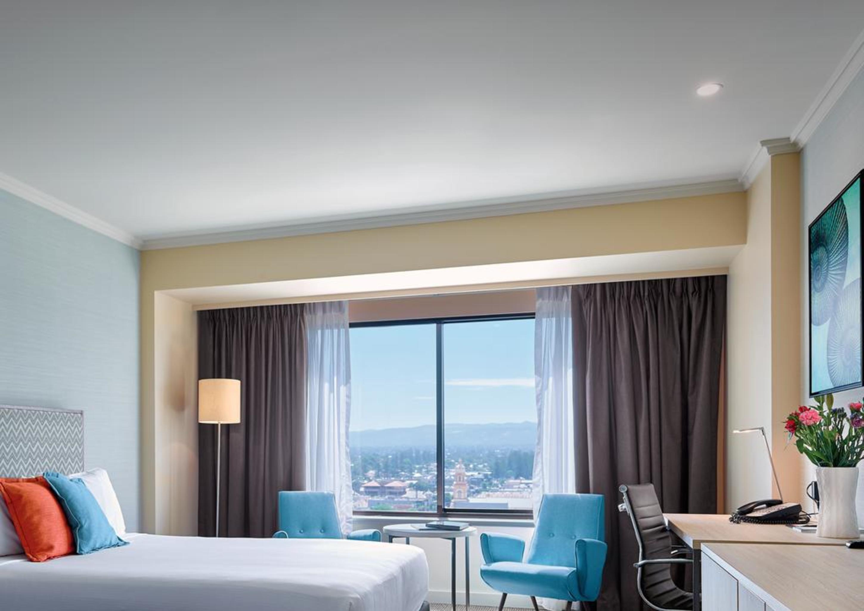 Stamford Grand Adelaide Hotel Glenelg Exterior photo The photo shows a modern hotel room featuring a queen-sized bed with white linens and colorful decorative pillows. There are two blue chairs beside a small table positioned near a large window that provides a view of the outside, with sheer and dark 
