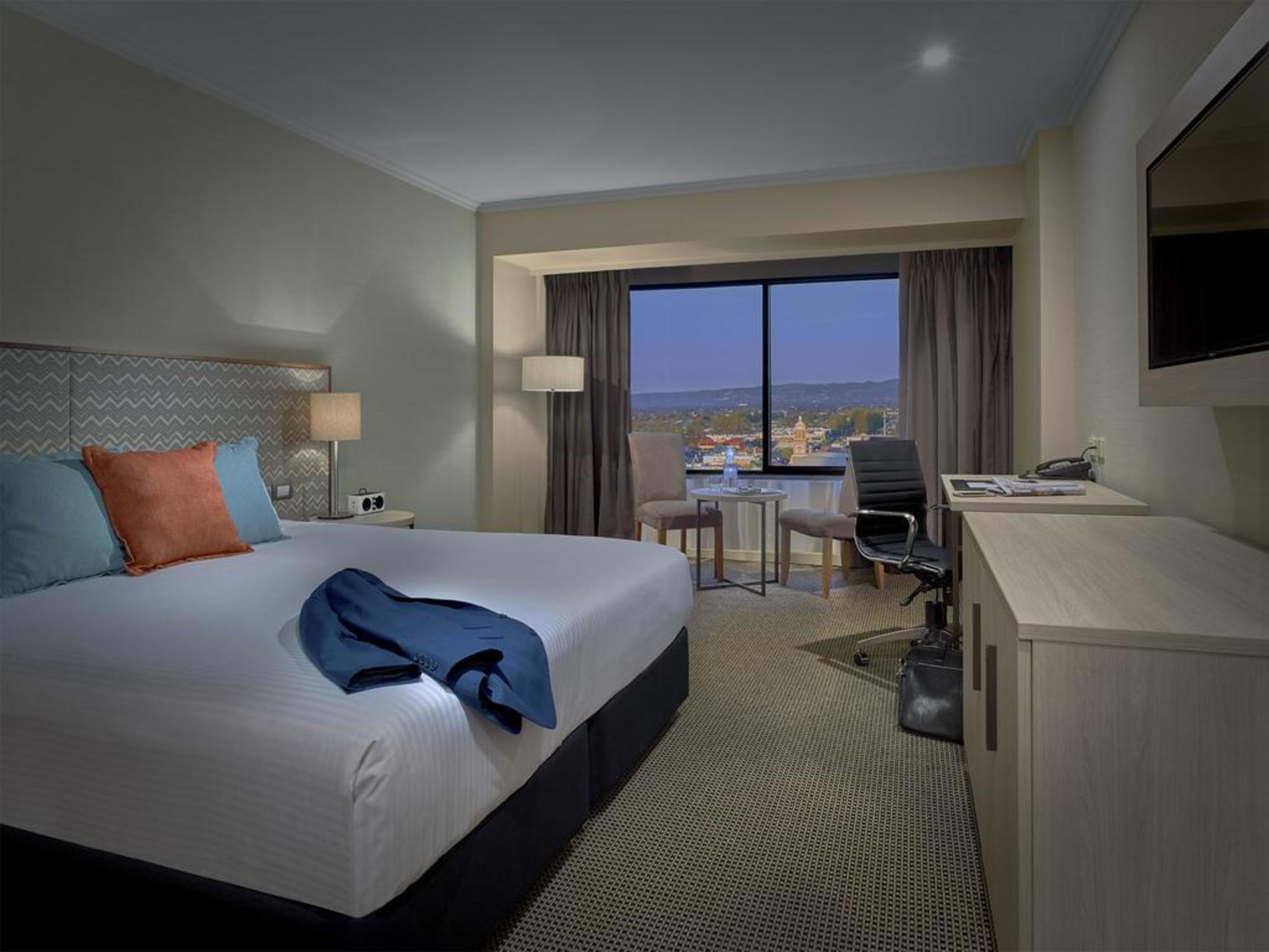 Stamford Grand Adelaide Hotel Glenelg Exterior photo The photo shows a modern hotel room. It features a large double bed with white bedding and decorative pillows in orange and blue. There's a nightstand with lamps on either side of the bed. A large window overlooks a view outside, and there's a desk a