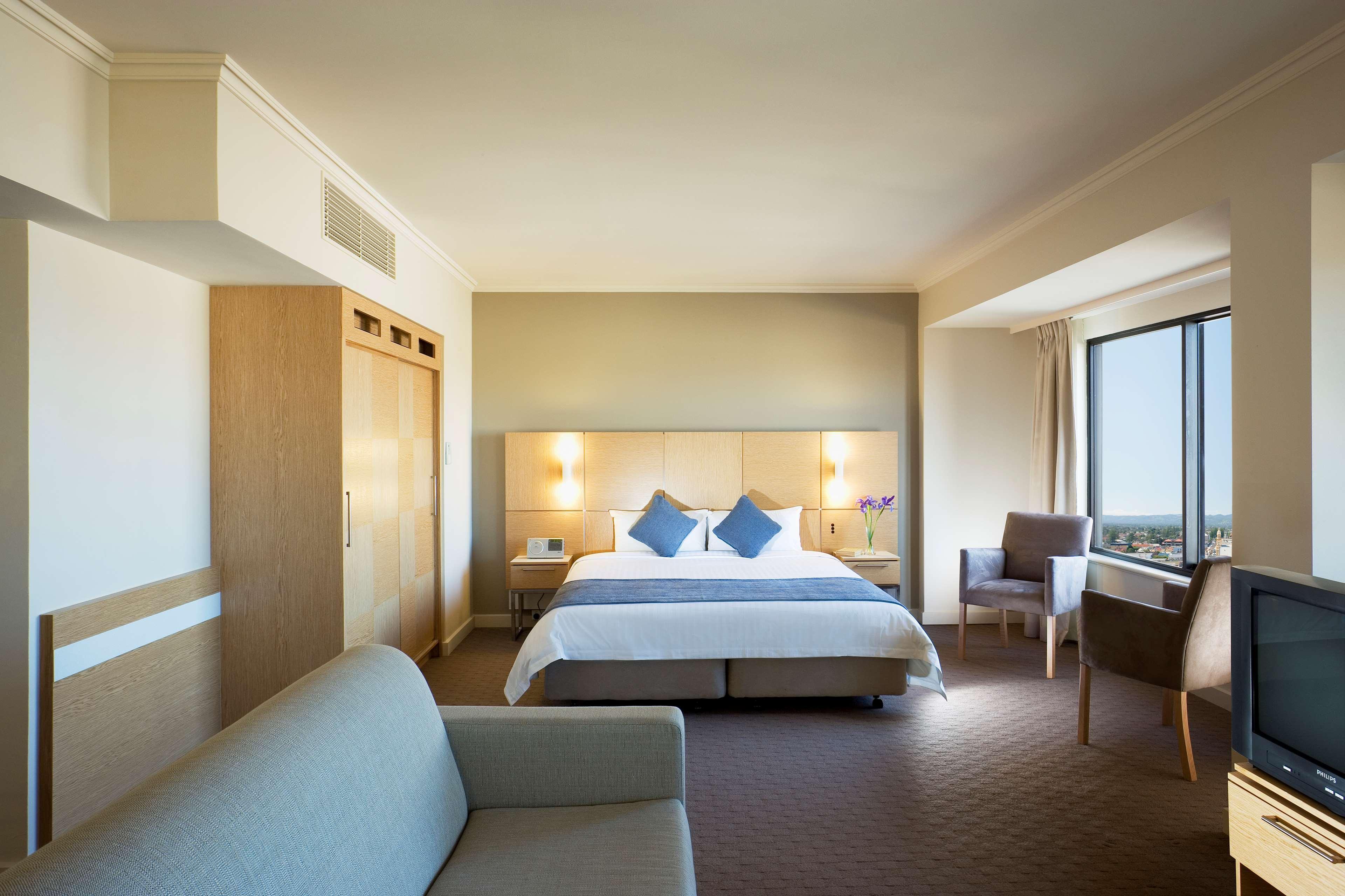 Stamford Grand Adelaide Hotel Glenelg Room photo The photo shows a modern hotel room featuring a large bed with two blue decorative pillows. There are bedside lamps on both sides of the headboard. To the left, there's a wooden wardrobe and a small table with a vase and a few amenities. On the right