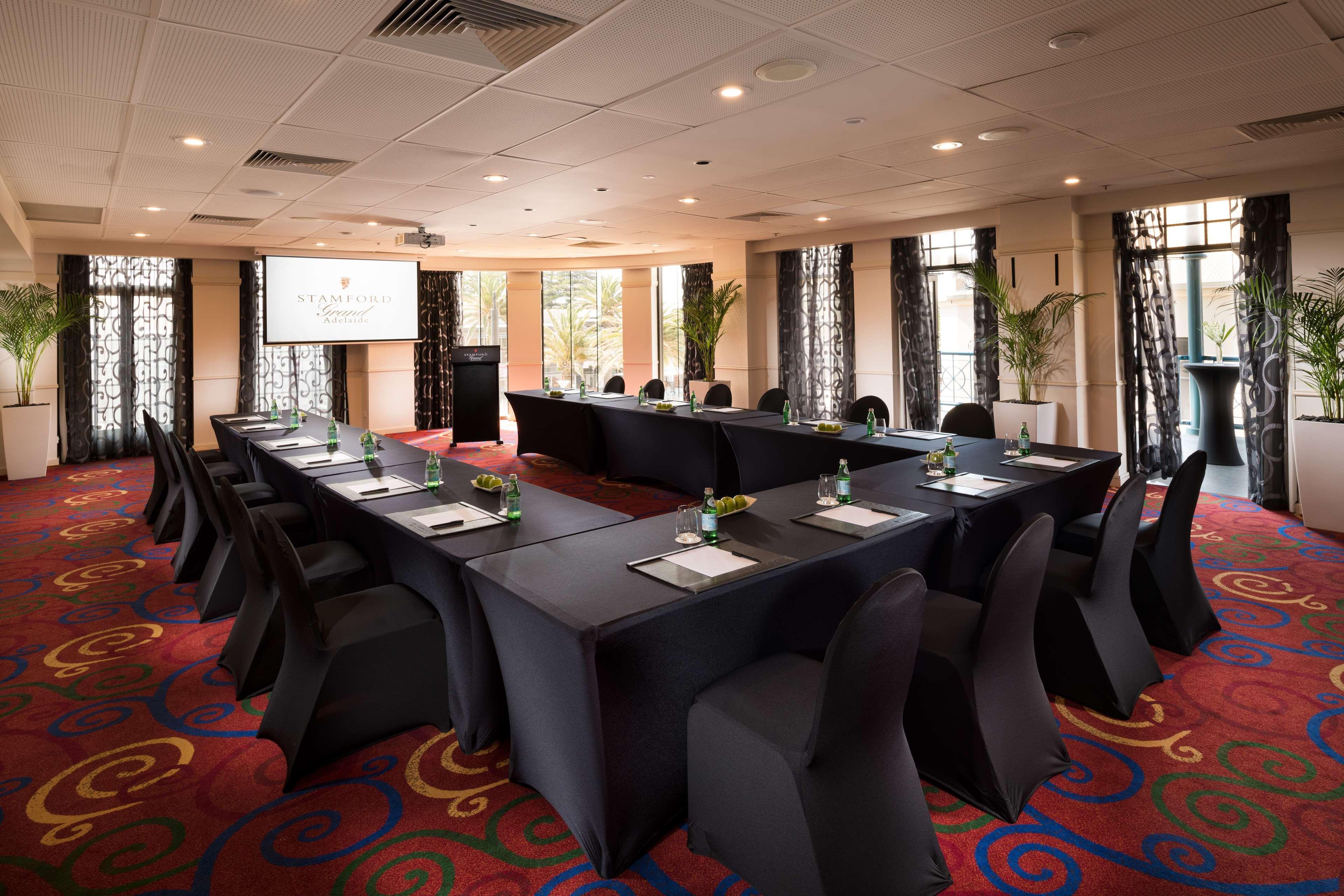 Stamford Grand Adelaide Hotel Glenelg Exterior photo The photo shows a conference room set up for a meeting or presentation. There are several tables arranged in a U-shape, covered with black tablecloths. Each table has a notepad and a small bottle of water on it. The chairs around the tables are also 