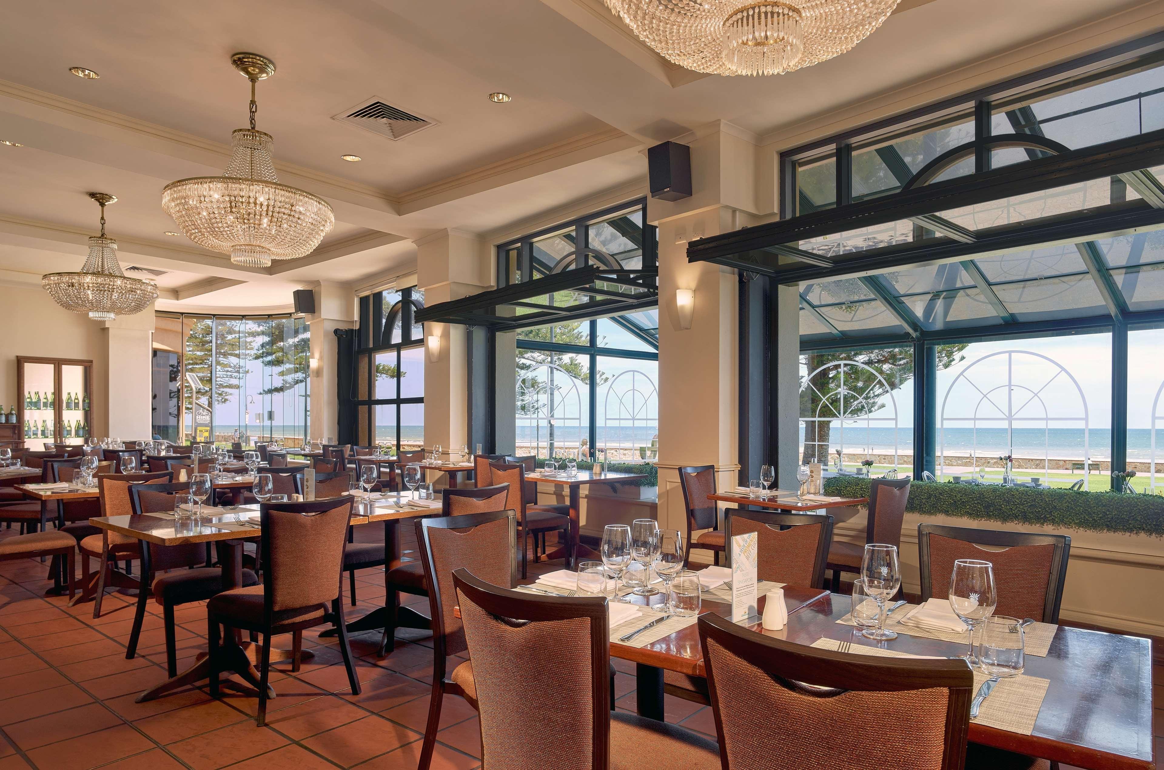 Stamford Grand Adelaide Hotel Glenelg Exterior photo The photo shows the interior of a restaurant. It features several wooden tables set with glasses and utensils, suggesting it's ready for diners. The decor includes elegant chandeliers hanging from the ceiling, adding a touch of sophistication. Large 