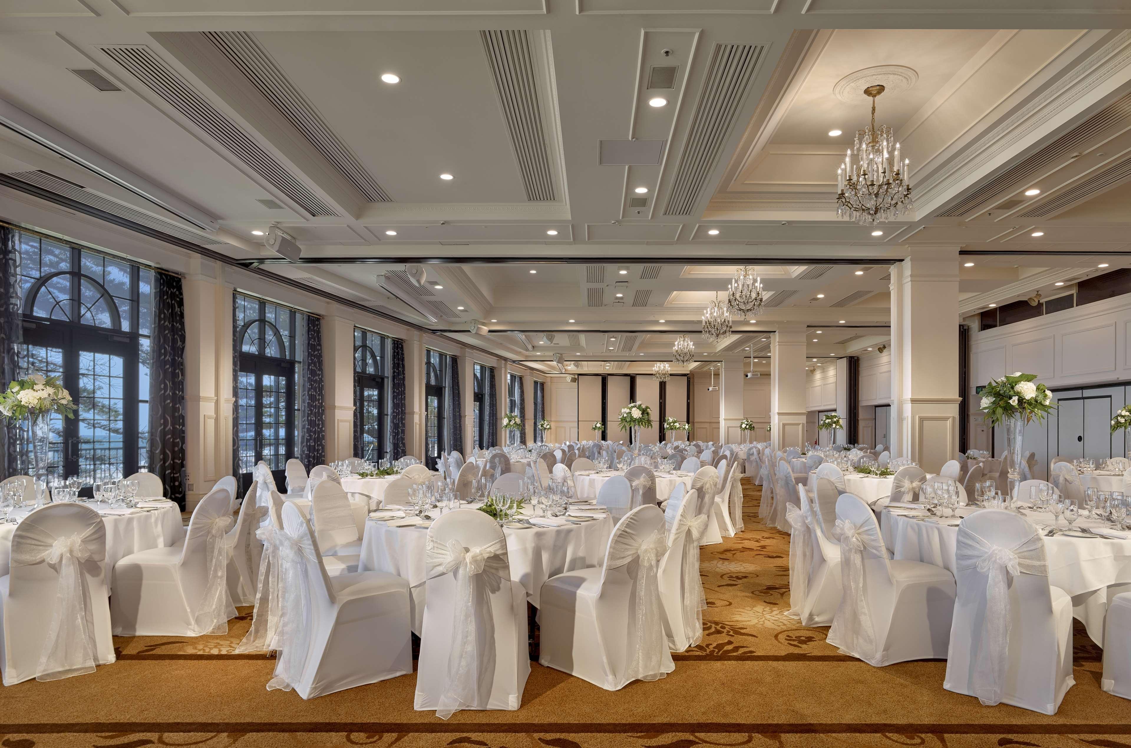 Stamford Grand Adelaide Hotel Glenelg Exterior photo The photo depicts an elegantly arranged banquet hall or dining area. The space features multiple round tables, each set with white tablecloths and decorated with floral centerpieces. The chairs are covered in white fabric, contributing to a formal at