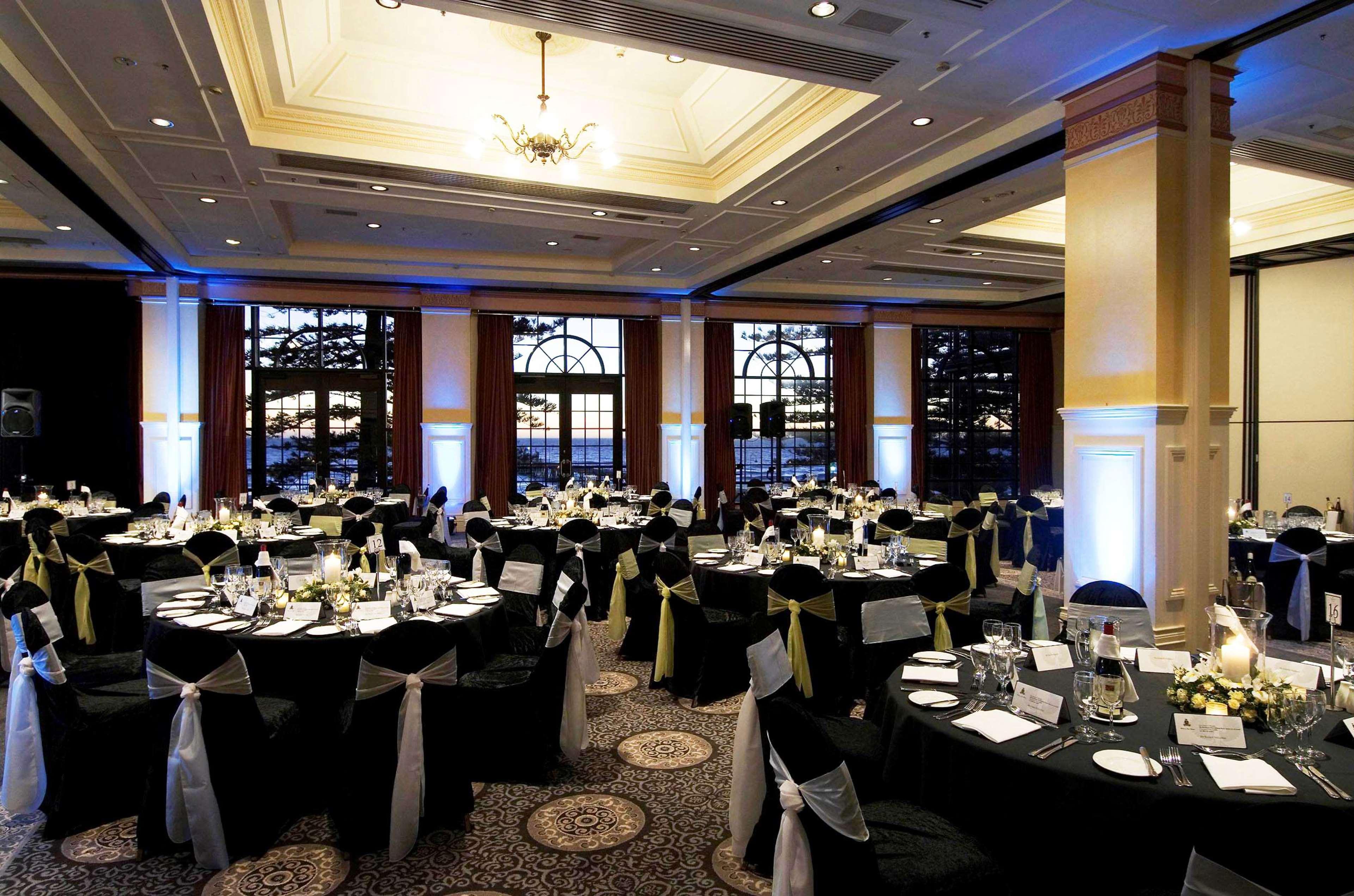 Stamford Grand Adelaide Hotel Glenelg Restaurant photo The photo depicts an elegantly arranged banquet hall set for an event. The room features round tables covered with black tablecloths, each adorned with white tableware and floral centerpieces. Chairs are draped in white fabric with decorative sashes 