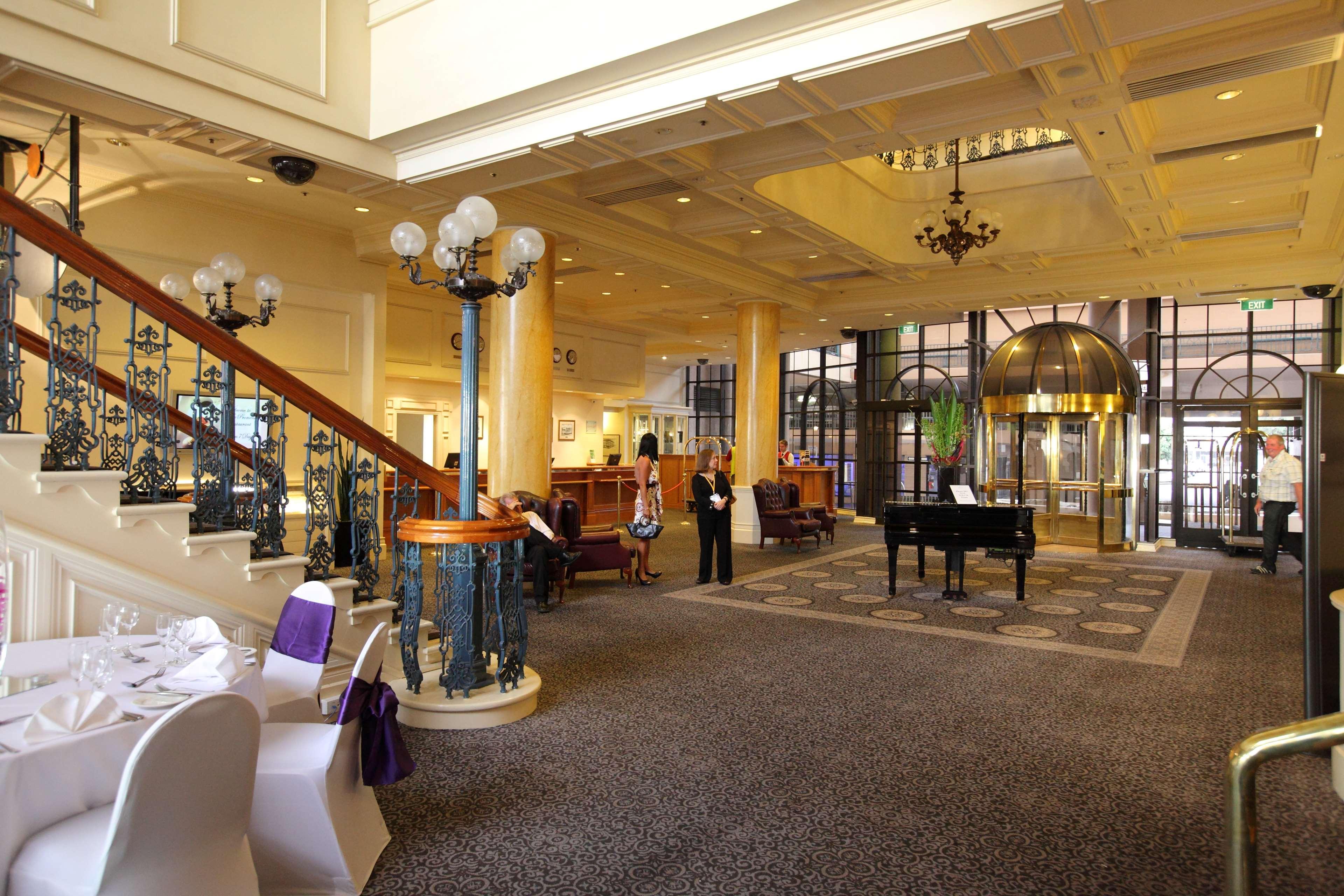 Stamford Grand Adelaide Hotel Glenelg Interior photo The photo shows a luxurious interior of a hotel or a restaurant lobby. There is an elegant staircase with ornate railings leading to an upper level. The space features high ceilings with decorative chandeliers and ample natural light coming through l