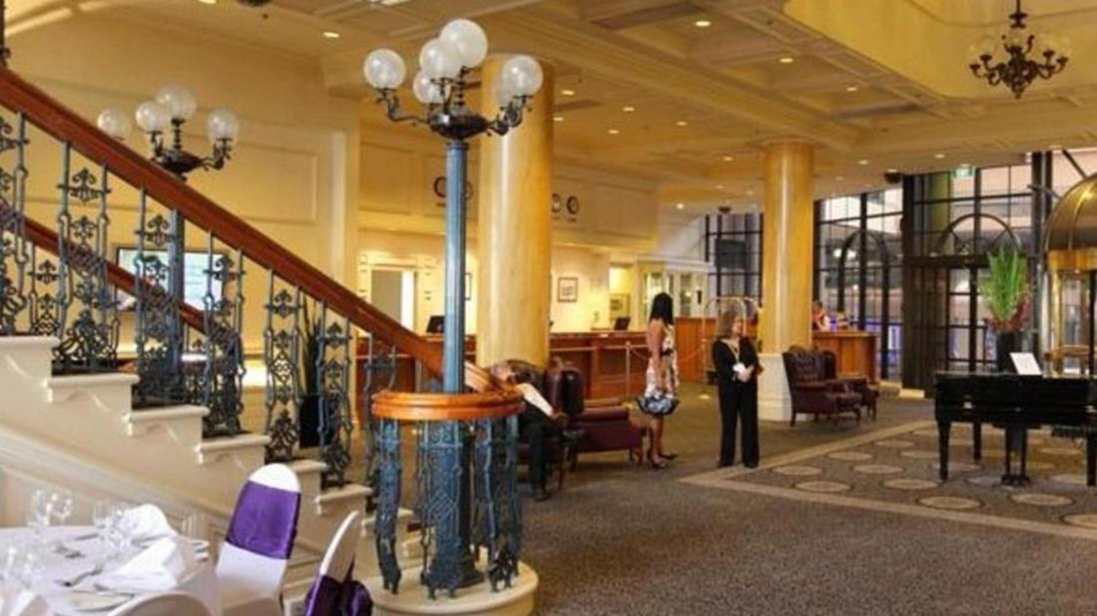 Stamford Grand Adelaide Hotel Glenelg Exterior photo The photo shows the interior of a hotel lobby. It features a spacious area with a high ceiling and elegant decor. There is a staircase with a decorative railing leading to an upper level. In the foreground, some guests and staff are present, and ther