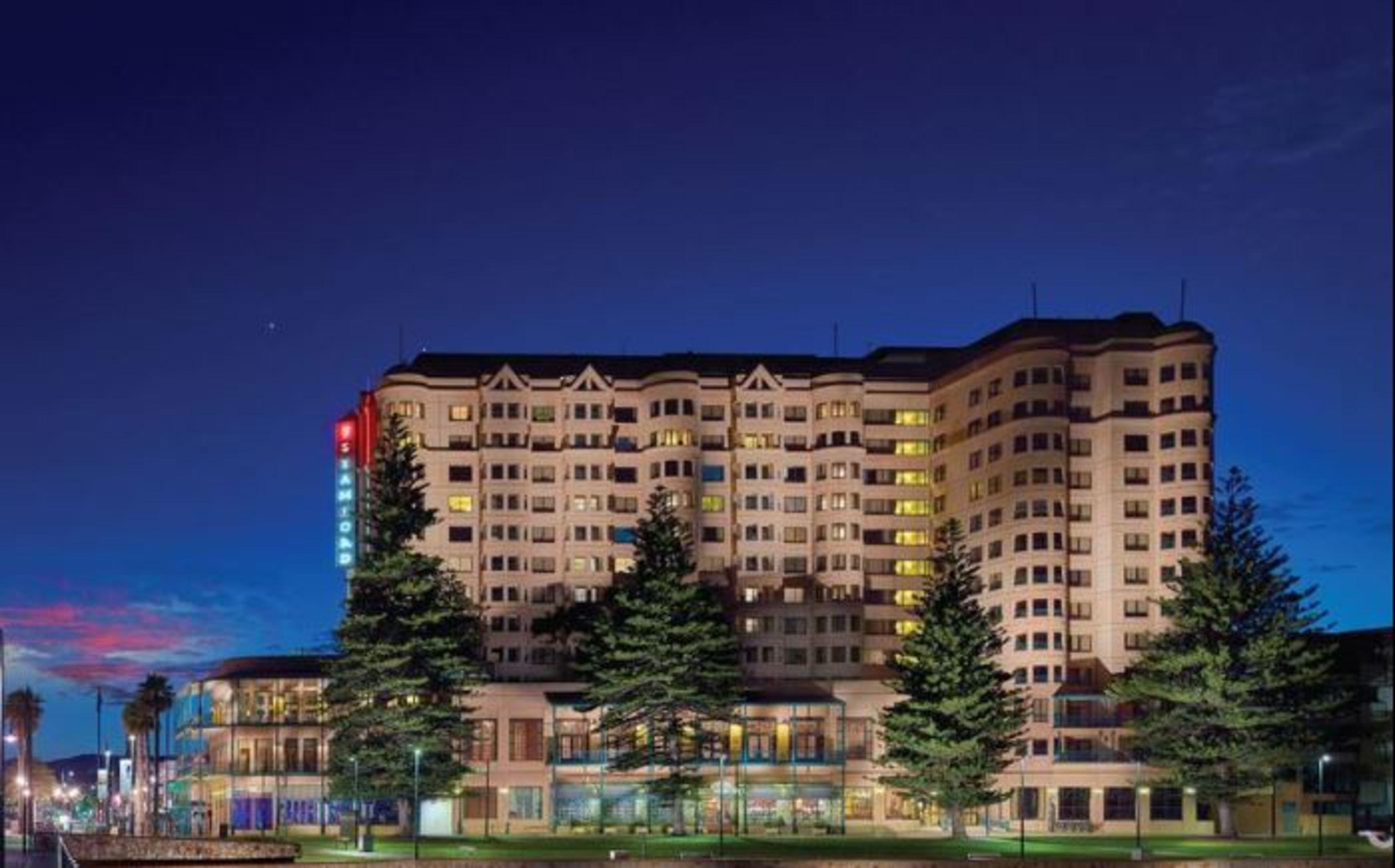 Stamford Grand Adelaide Hotel Glenelg Exterior photo The photo displays a multi-story hotel building at dusk, illuminated by various lights. The architecture features a mix of modern and traditional design elements, with numerous windows showcasing a warm glow. Surrounding the hotel are several tall tr