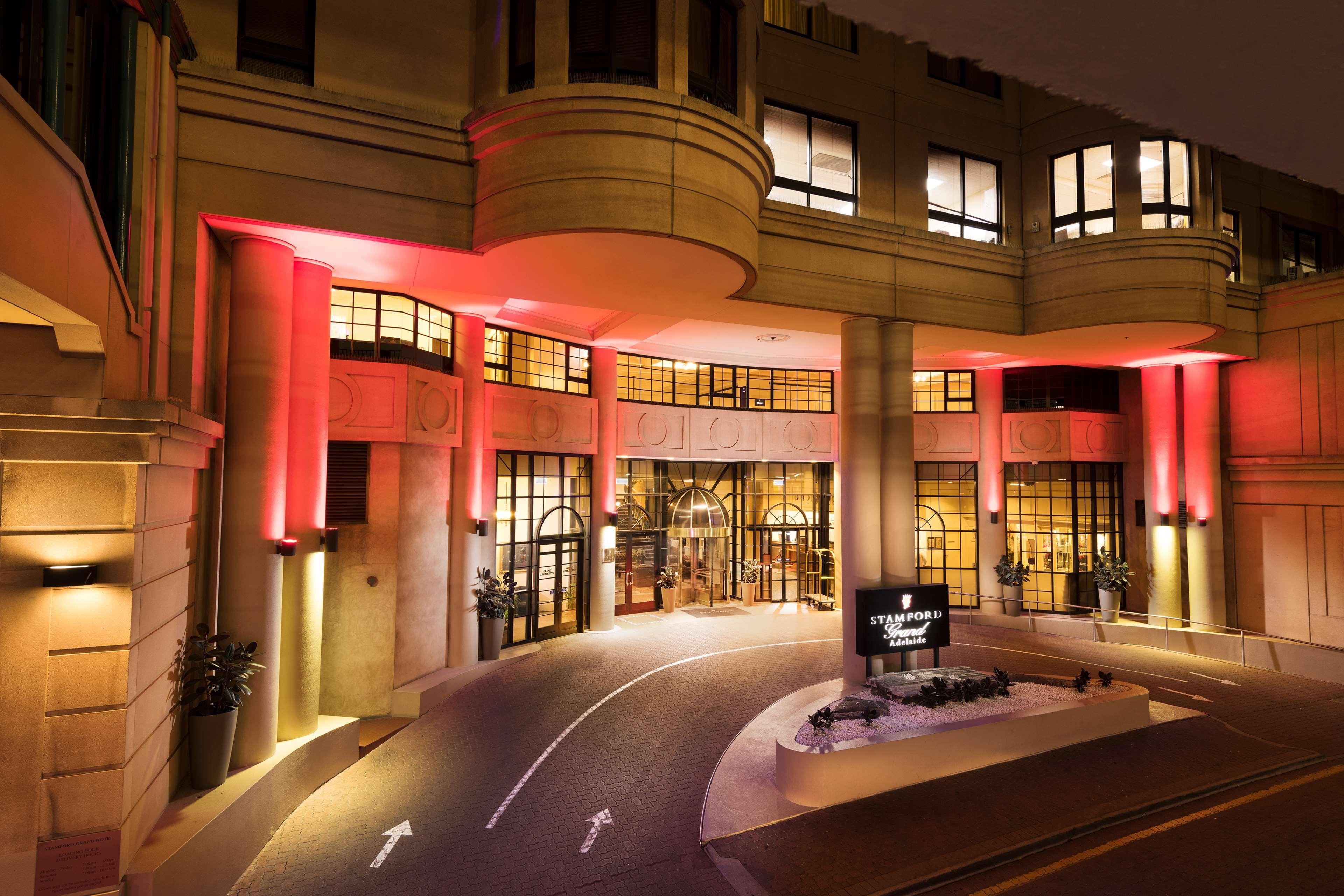 Stamford Grand Adelaide Hotel Glenelg Exterior photo The photo shows the entrance of a hotel or luxury building at night. The façade features a modern architectural style with large windows and an elegant entrance. The area is illuminated with red lighting, creating an inviting atmosphere. There appear
