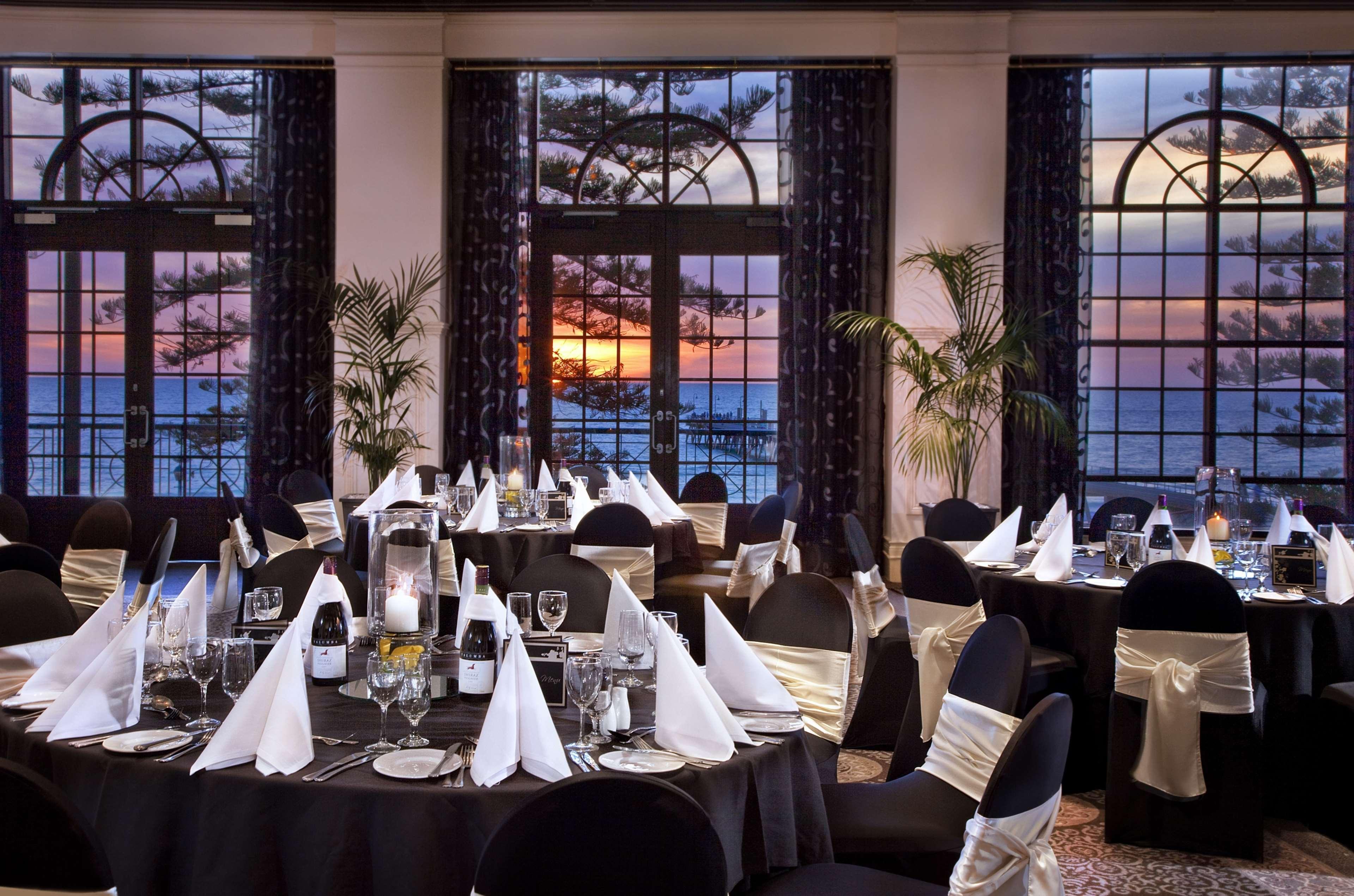 Stamford Grand Adelaide Hotel Glenelg Restaurant photo The photo shows an elegantly decorated dining room with a beautiful view. The tables are set with black tablecloths and white napkins folded in a triangular shape. There are glassware and silverware neatly arranged at each setting. The room is adorne