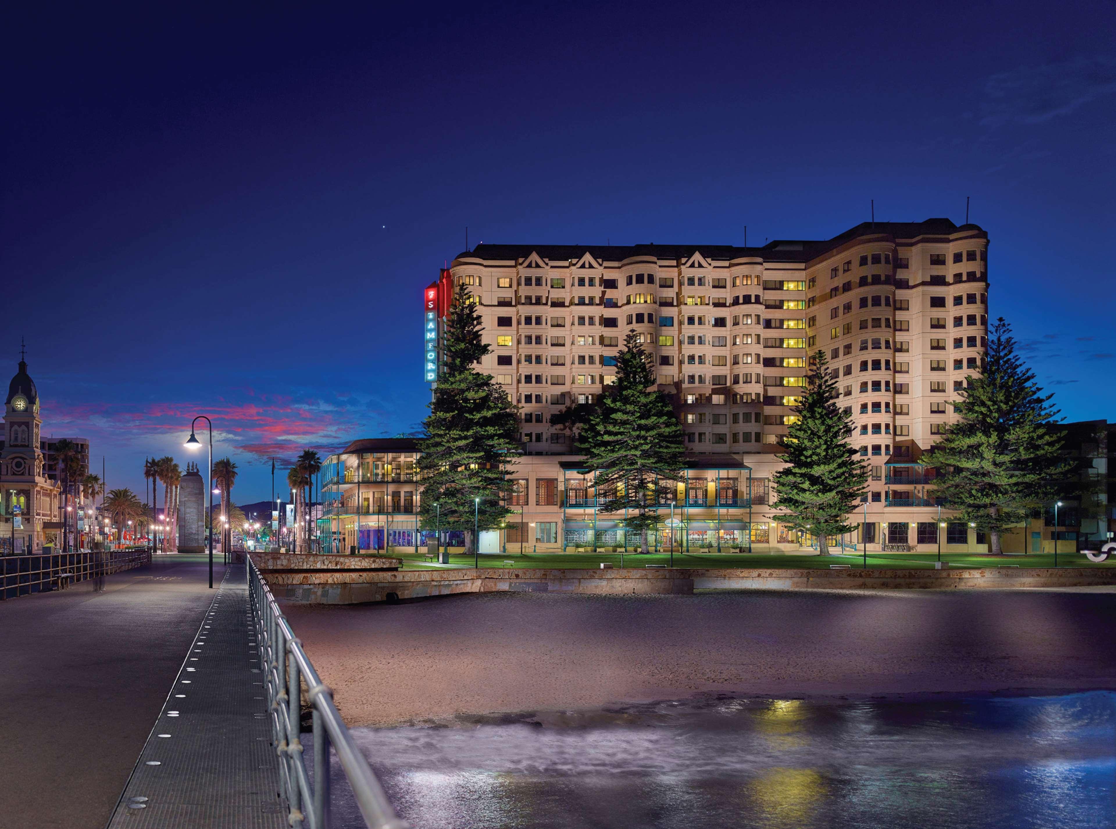 Stamford Grand Adelaide Hotel Glenelg Exterior photo The photo shows a waterfront scene at dusk, featuring a large building lit up against a darkening sky. The architecture appears modern and prominent, with several windows illuminated. In the foreground, there is a walkway or pier with fencing along t