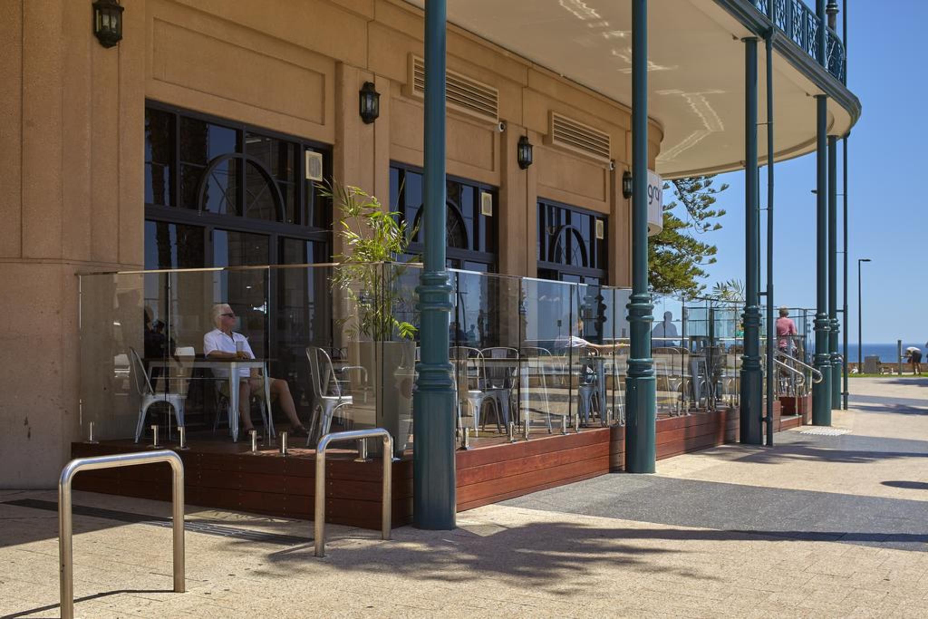 Stamford Grand Adelaide Hotel Glenelg Exterior photo The photo shows the exterior of a cafe or restaurant with a large outdoor seating area. There are glass barriers around the seating, providing a view of the outside while maintaining a sense of enclosure. The building has a modern architectural style