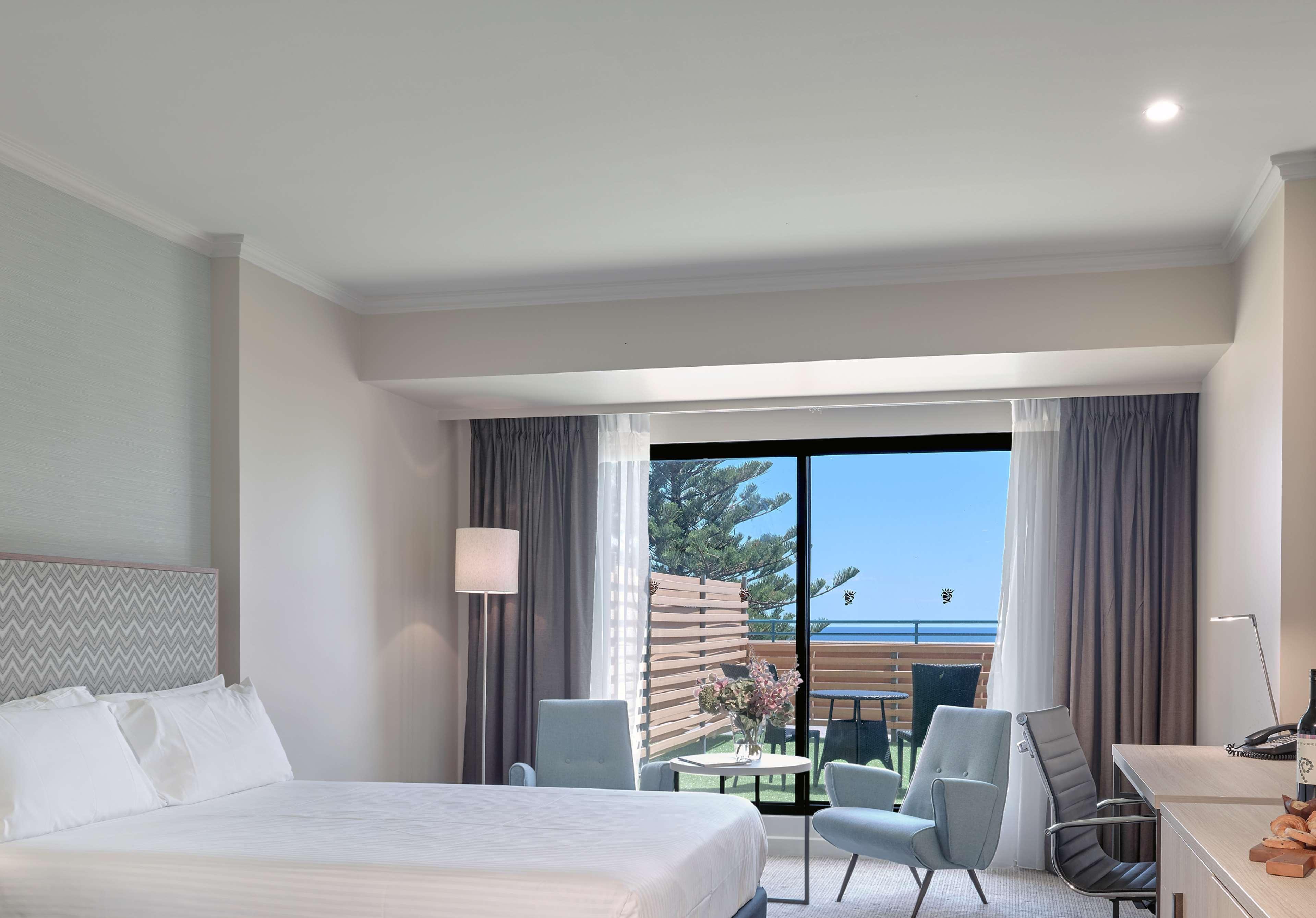 Stamford Grand Adelaide Hotel Glenelg Exterior photo The photo shows a modern hotel room featuring a large bed with white bedding. There are two armchairs positioned near a window that offers a view of an outdoor area, possibly a balcony or patio with greenery. The room has light-colored walls and curt