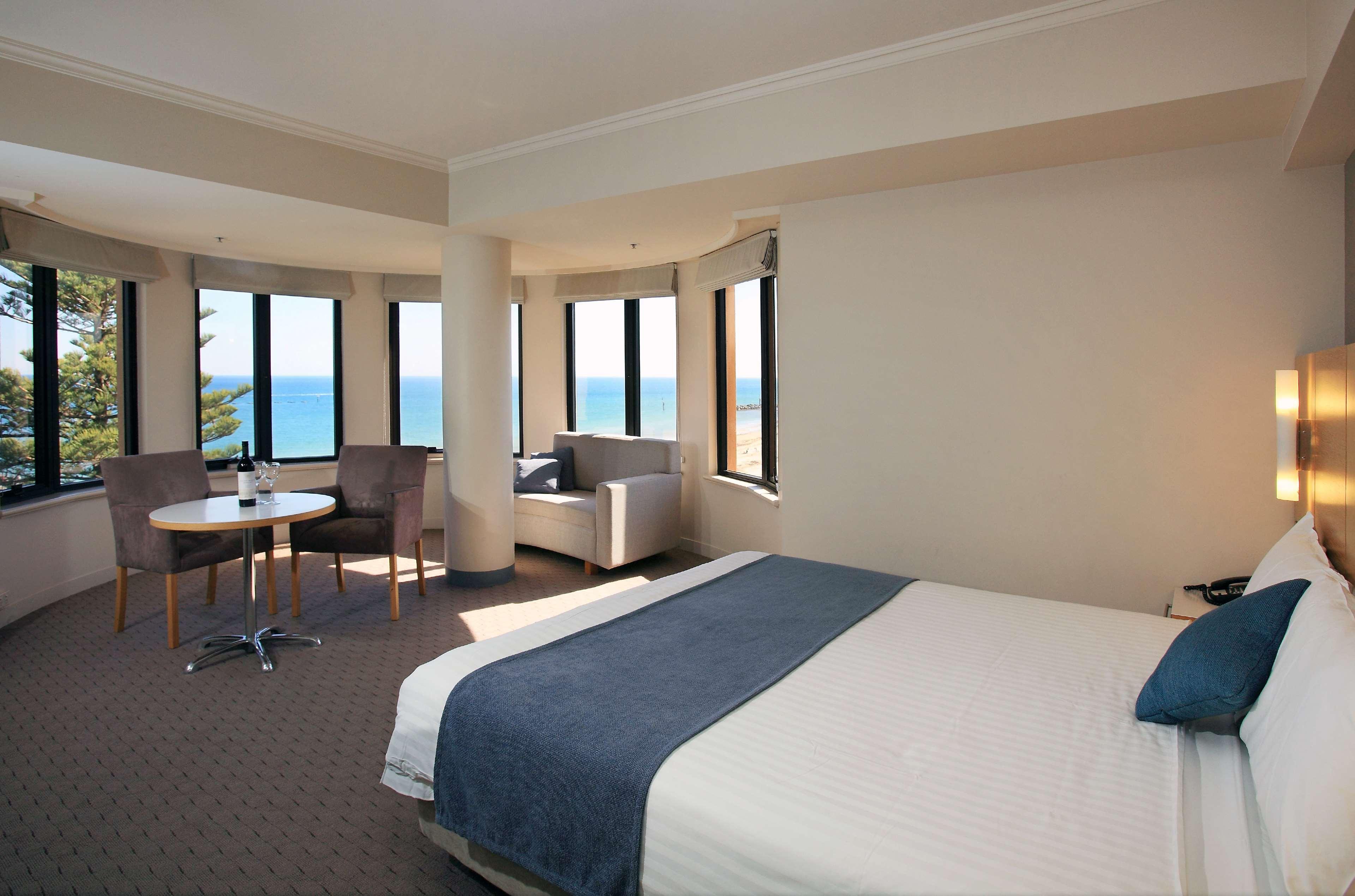 Stamford Grand Adelaide Hotel Glenelg Exterior photo The photo shows a well-lit hotel room featuring large windows that offer a view of the ocean. The room is arranged with a comfortable bed covered in white linens and a navy blue throw blanket. There is a small table and chairs set up beside the windo