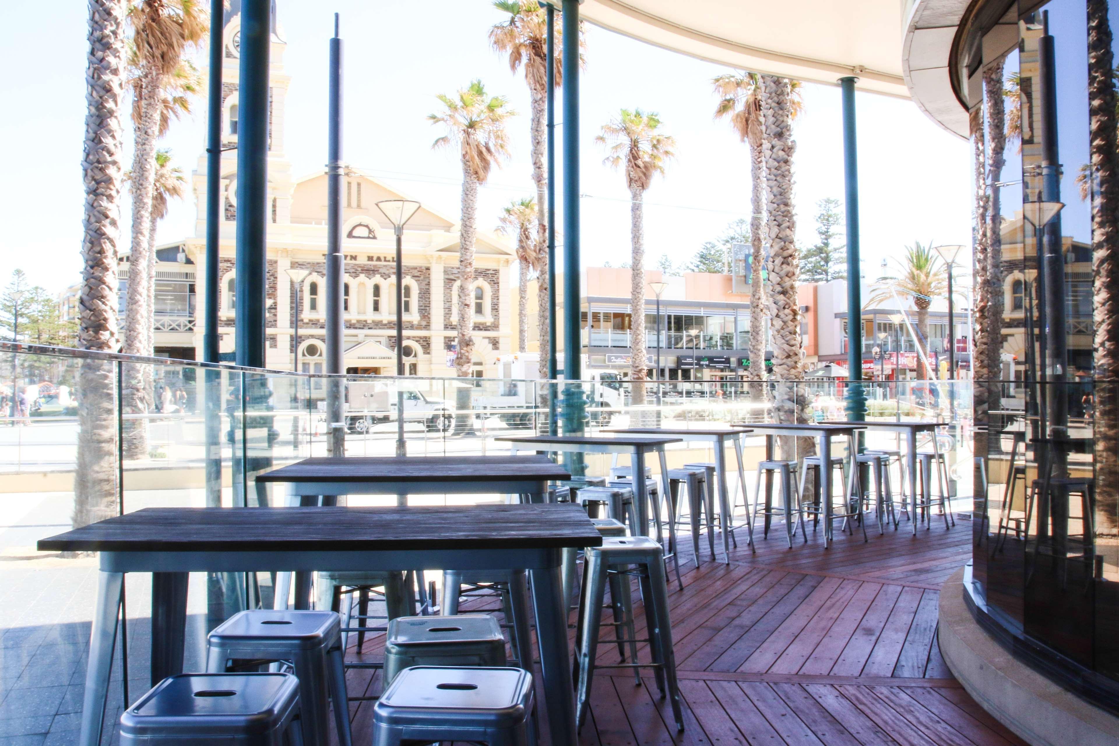 Stamford Grand Adelaide Hotel Glenelg Exterior photo The photo shows an outdoor seating area of a café or restaurant. It features metal tables and stools arranged on a wooden deck. The setting has large glass panels, providing an open and airy atmosphere. In the background, palm trees and a building wi