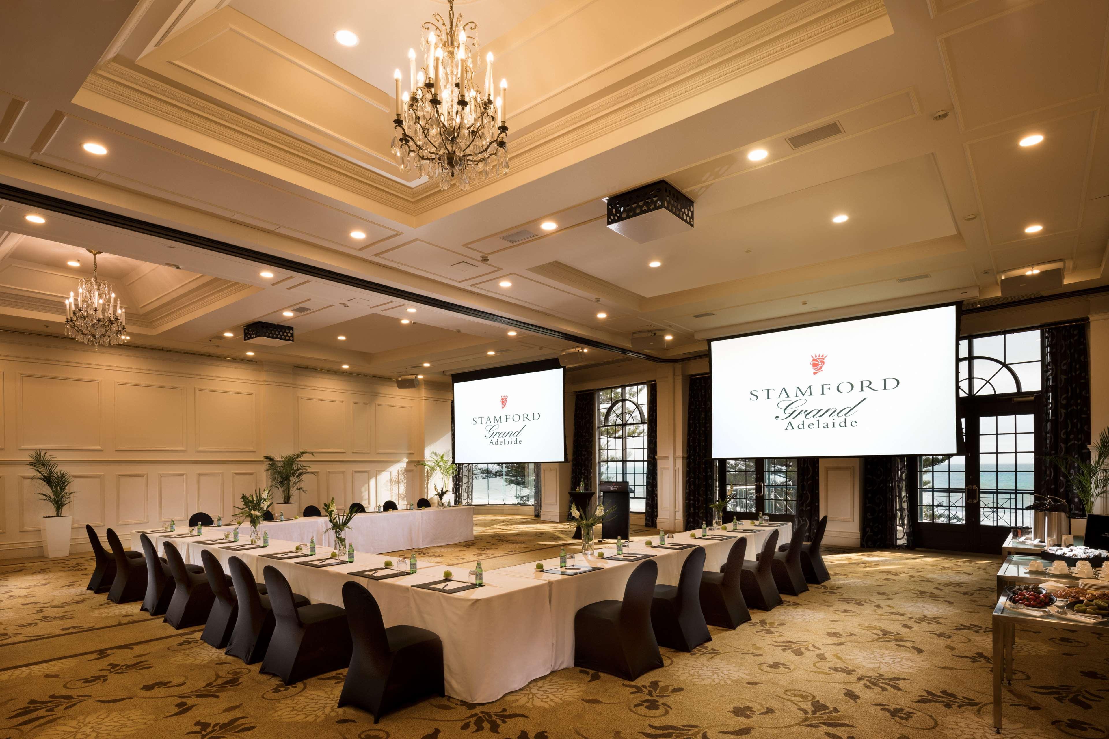 Stamford Grand Adelaide Hotel Glenelg Exterior photo The photo shows a spacious and elegantly designed conference room. The room features a long table set for a meeting, surrounded by stylish chairs. Two large screens are positioned at the front, displaying the logo "Stamford Grand Melbourne." The ceil