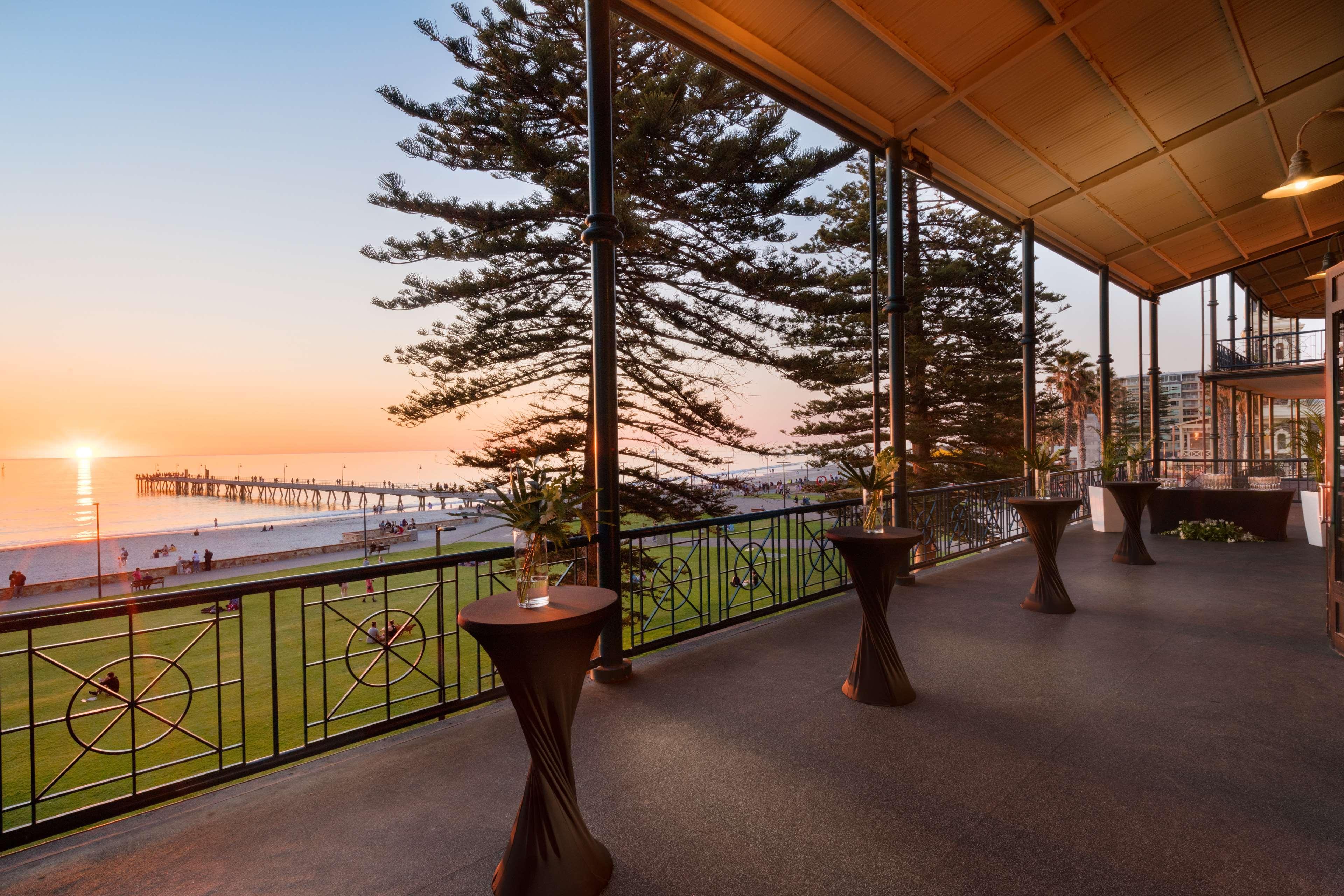 Stamford Grand Adelaide Hotel Glenelg Exterior photo The photo shows a spacious indoor space with large windows overlooking a beach at sunset. The scene features a pier extending into the ocean and a horizon painted in warm colors from the setting sun. In the foreground, there are elegant tables with d