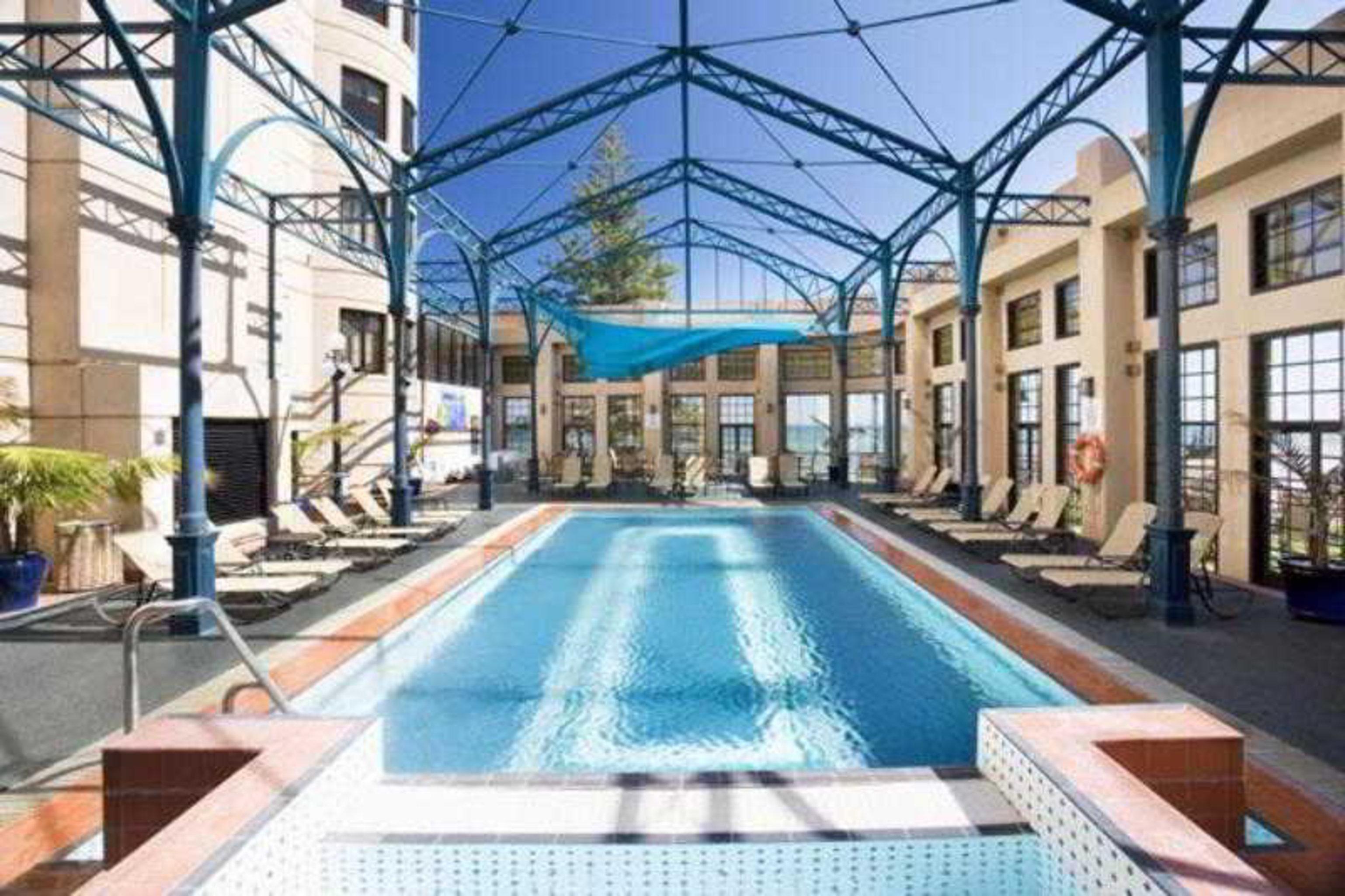 Stamford Grand Adelaide Hotel Glenelg Facilities photo The photo shows an indoor swimming pool area. The pool is rectangular and features a blue cover above, supported by a metal frame. Surrounding the pool are several lounge chairs, and the space is well-lit with large windows allowing natural light to 