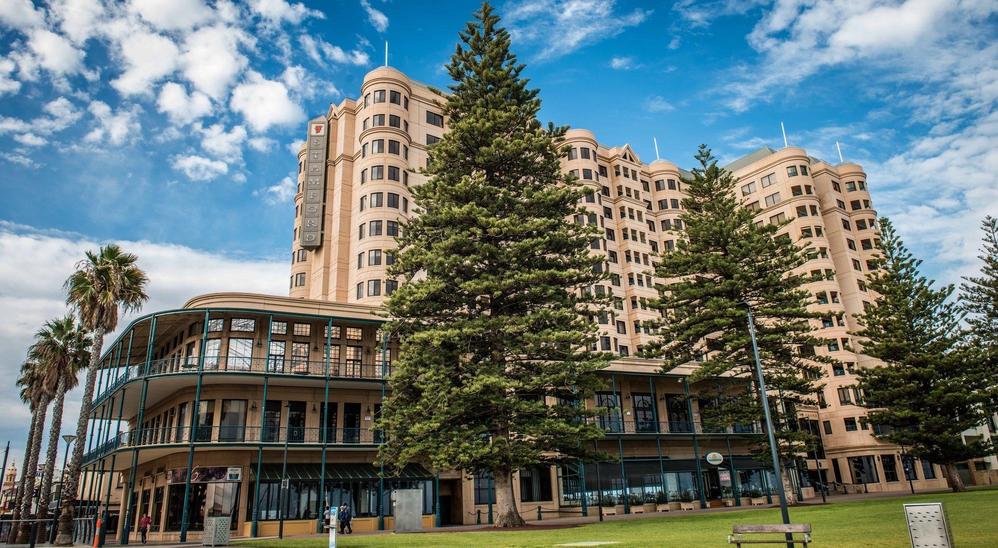 Stamford Grand Adelaide Hotel Glenelg Exterior photo The image shows a large building with a distinctive architectural style, characterized by a tan or beige color. The structure appears to have multiple floors and rounded edges. In front of the building, there are several tall green trees, which add t