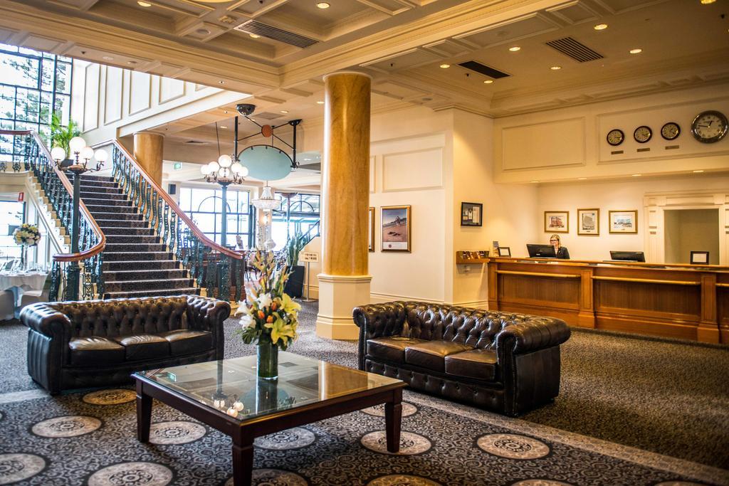 Stamford Grand Adelaide Hotel Glenelg Exterior photo The photo depicts a hotel lobby featuring a spacious and elegant design. There are two dark leather sofas arranged in front of a glass coffee table, adorned with a floral centerpiece. The lobby has a refined atmosphere, highlighted by large windows t
