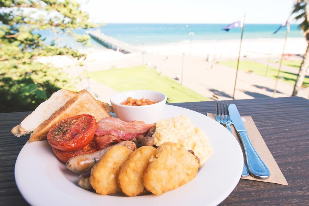 Stamford Grand Adelaide Hotel Glenelg Exterior photo The photo shows a plate of breakfast food set against a beachside backdrop. The plate includes several items: crispy bacon, a grilled tomato, sausage links, golden hash browns, and a small serving of baked beans. There is also a piece of toast on the